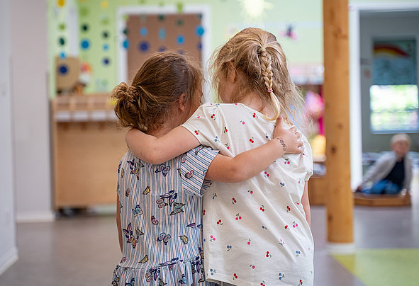 Zwei Mädchen umarmen sich von hinten in einem hellen Kitaraum; bunte Wände und Spielmaterialien im unscharfen Hintergrund.
