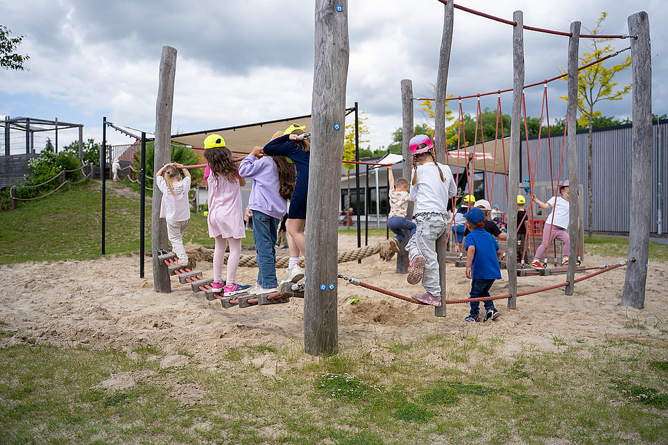 Mehrere Kinder spielen auf einem hölzernen Kletterparcours im Sand mit Balancierseilen, Netz und Pfosten; Außenspielbereich unter bewölktem Himmel.