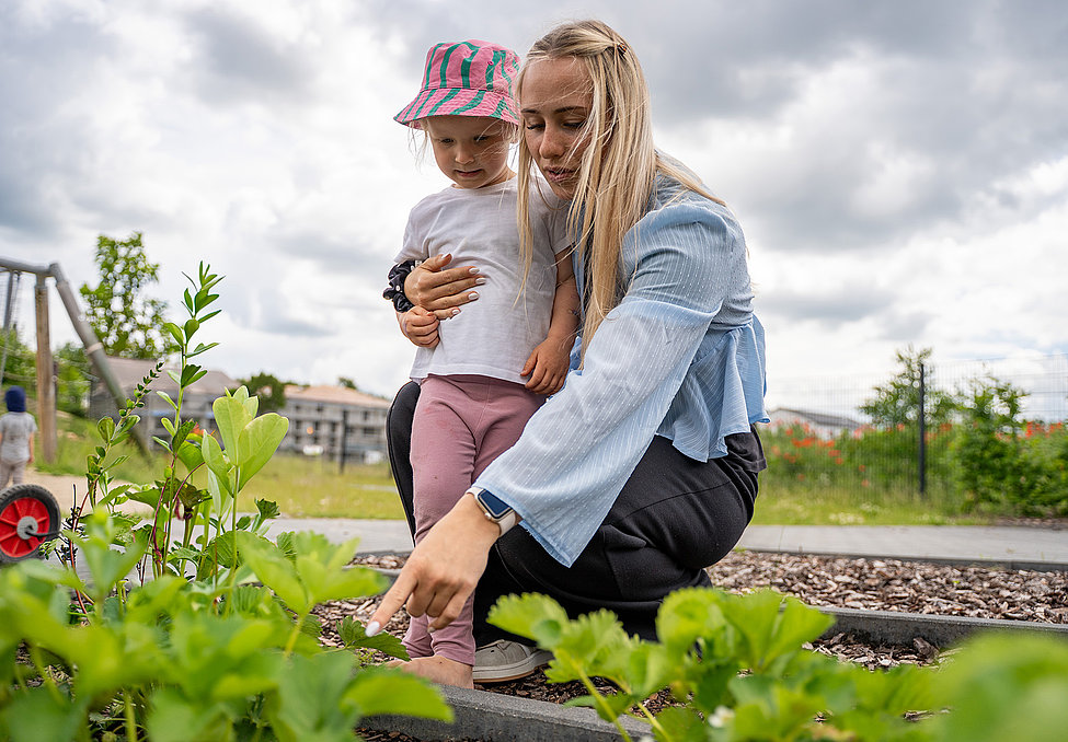 Betreuerin hockt im Beet, zeigt Pflanzen und hält ein Kleinkind im Arm; Spielplatz und bewölkter Himmel im Hintergrund.