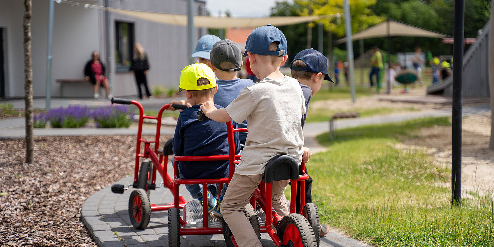 Vier Kinder sitzen auf einem roten Vierradwagen und fahren einen gepflasterten Weg im Kita-Außenbereich; Sonnenschutz und Gebäude im Hintergrund.